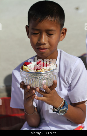 A young Thai boy makes a prayer before scattering a bowl of rose petals in front of Buddhist monks. Stock Photo