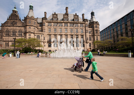 A mother and child both dressed in green sweatshirts, mother with a buggy, walk in front of the fountain and Sheffield Town Hall Stock Photo