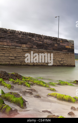old pier with lantern with in front seaweed covered rocks long exposure gives seawater and clouds a milky look Stock Photo