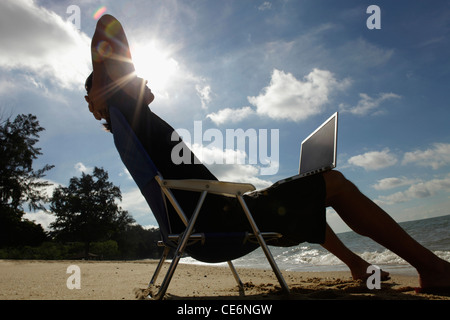 Man sitting in beach chair with laptop, sun burst behind his head Stock Photo
