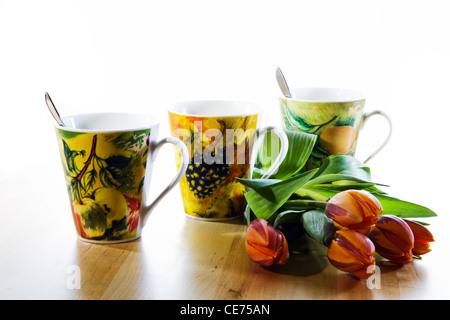 Colorful still-life with three decorated mugs, orange tulips and white background on wooden table Stock Photo