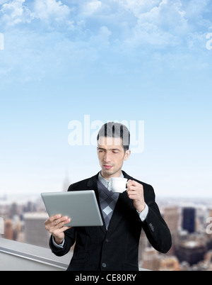 Young businessman with a cup of coffee uses a digital tablet on the roof of business center. Blurred cityscape with skyscrapers Stock Photo