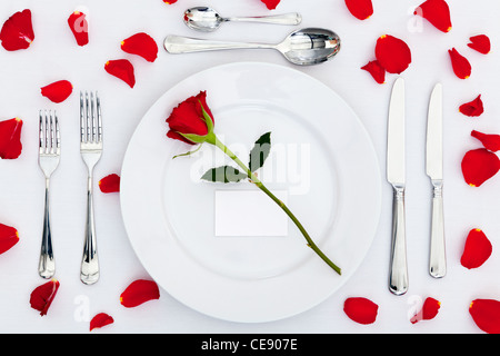 Photo of a table place setting with a red rose and blank card on the plate plus rose petals on the tablecloth Stock Photo