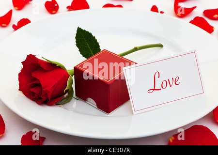 Photo of a red rose on a plate with a box for an engagement ring and a card with the word Love Stock Photo