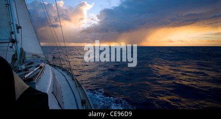 A sailboat on an ocean passage heads toward the sun and a small rain squall. Stock Photo