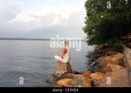 A westerner staying on retreat at a Buddhist temple meditates infront of a large lake Stock Photo