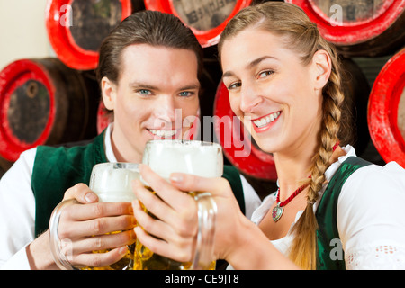 Young couple, man and woman, in traditional Bavarian Tracht drinking beer in a brewery in front of beer barrels Stock Photo