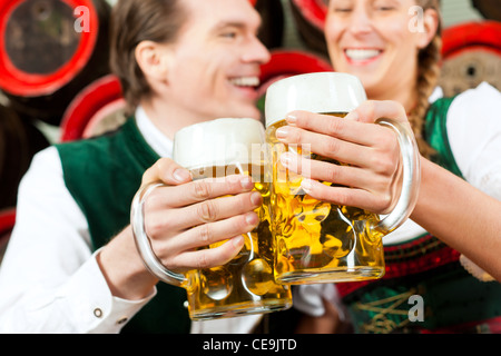 Young couple, man and woman, in traditional Bavarian Tracht drinking beer in a brewery in front of beer barrels Stock Photo