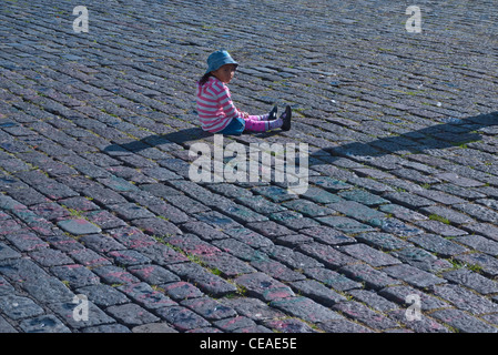 A 4-5 year old girl sits on the granite paver stones of a plaza in Quito, Ecuador with only the shadow of another person visible Stock Photo
