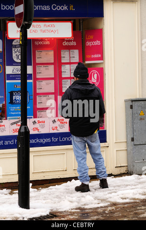 A man standing on a snowy high street looking at holidays in a travel agents window display Stock Photo