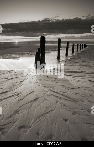 The sun sets behind a row of old wooden groynes on a sandy beach in Brancaster, Norfolk Stock Photo