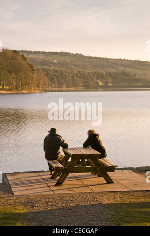 Couple (man & woman) sitting relaxing, on picnic table bench looking out over water on sunny day - scenic Swinsty reservoir, Yorkshire, England, UK. Stock Photo