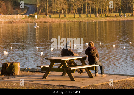 Couple (man & woman) sitting relaxing, on picnic table bench looking out over water on sunny day - scenic Swinsty reservoir, Yorkshire, England, UK. Stock Photo