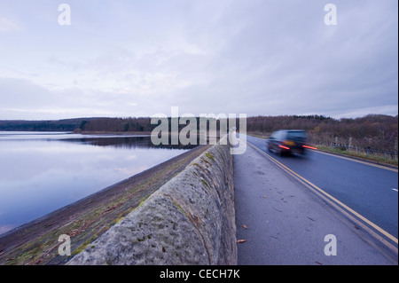 Car (4X4 vehicle + tail lights on) driving on road that crosses dam embankment by Fewston Reservoir (trees reflected in calm water) - Yorkshire, UK.