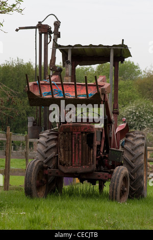 An old red tractor sitting in a field with worn tyres and its hay scoop raised Stock Photo