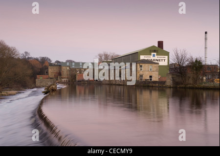 Flowing water of River Wharfe cascading over weir under pink sunset sky, historic Garnett's paper mill beyond - Otley, West Yorkshire, England, UK. Stock Photo