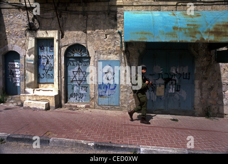 An armed Israeli Soldier walks past sealed Palestinian shops which are sprayed with the Jewish symbol Star of David and inflammatory phrases in Hebrew which reads 'Revenge', 'We will take Revenge' and 'Arabs Out' in Al-Shuhada Street which used to be the central wholesale market of the Hebron region and was closed to Palestinian merchants after violence in the Second Intifada in old city of Hebron West Bank Israel Stock Photo