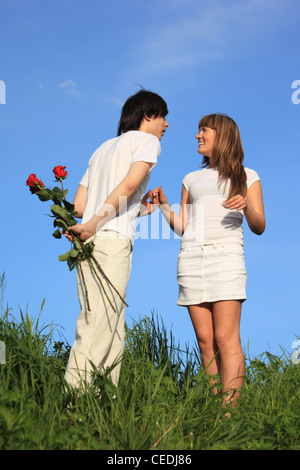 young pair stands on a grass,  guy holds behind back bouquet of roses Stock Photo