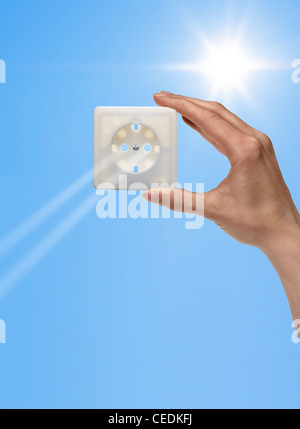 symbolic solar energy theme showing the sun and sky with human hand holding a electrical socket while sunbeams falling through Stock Photo
