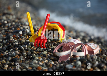 Yellow children's bucket with a scoop, a red rake and sandals on seacoast Stock Photo