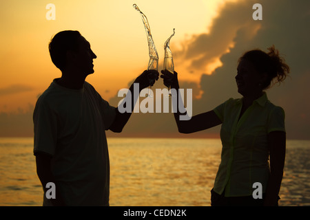 Man and woman clink glasses. Splashes of wine from glasses. Silhouettes against sea. Stock Photo