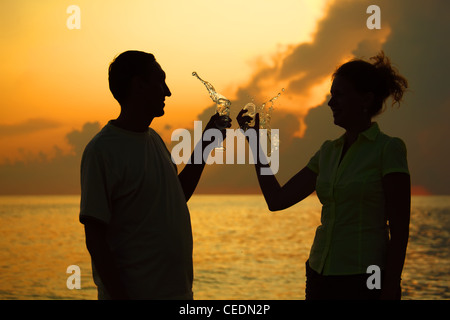 Man and woman clink glasses. Splashes of wine from glasses. Silhouettes against sea. Stock Photo