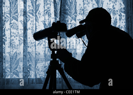 Man with binoculars and camera with long lens on tripod looking through net curtains. Photograph posed by model. Stock Photo