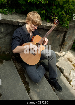 Young man sitting on old cement stairway, playing a classical guitar.