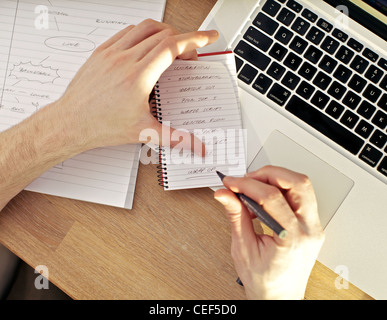Overhead view of a mans hands writing in a notebook balanced on a laptop keyboard. Stock Photo