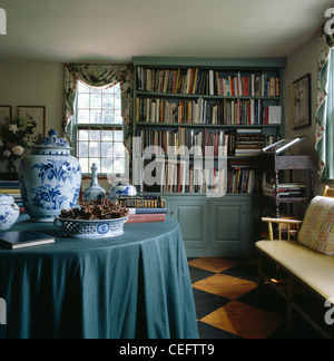 Sage-green cloth on table with blue+white china pots in country dining room with sage-green book-case and diamond-painted floor