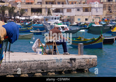 A man fishes from the harbour side at Marsaxlokk with the 'Luzzu' - traditional Maltese fishing boats - moored in the bay Stock Photo