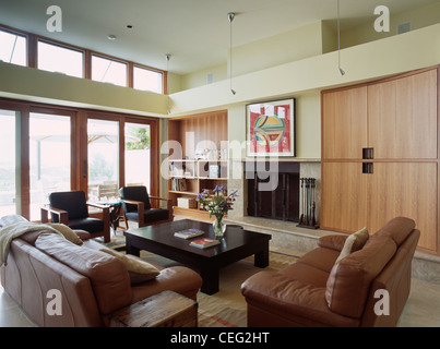 Brown leather sofas in modern double-height living room with large fitted storage cupboard beside fireplace Stock Photo