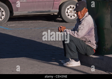 A 70 to 80 year old man, with a black baseball cap and white tennis shoes, sits on a curb in Latacunga, Ecuador. Stock Photo