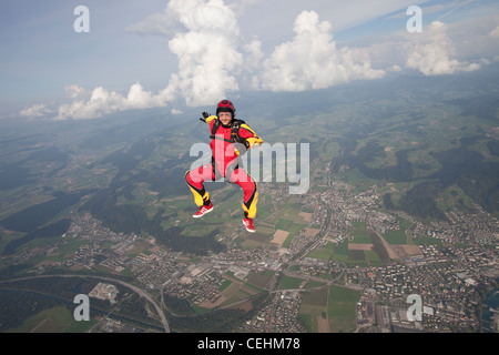 Skydiver girl is flying in sit position in free fall. She is flying free in the blue sky over a small town and green fields. Stock Photo