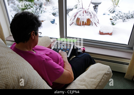 Senior mature pensioner woman holding Apple iPad digital tablet computer selecting apps by window sitting in armchair winter snow outside England UK Stock Photo