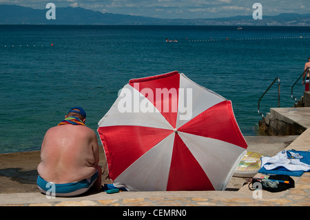 Man sits by the water on a Croatia beach next to his red and white umbrella. Stock Photo