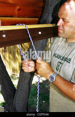 FORBuild a Tree House Step Male tying the rope to the floof the tree house to create a hanging rope ladder Stock Photo