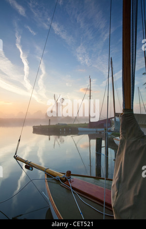 View over the bow of a sailing boat on the River Thurne through dawn mist towards Thurne drainage mill on the Norfolk Broads. Stock Photo