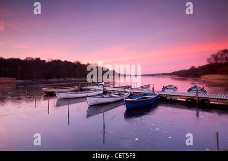 Sunrise over rowing boats tied to a jetty on the frozen water of Ormesby Broads in the Broads National Park in Norfolk. Stock Photo