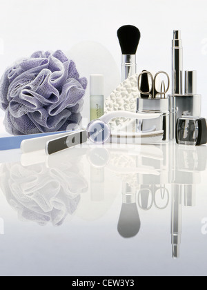 Assorted beauty and grooming tools including makeup brush, scissors, nail care items and bath sponge arranged on a white reflective surface. Stock Photo