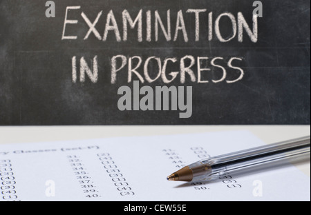 Close up on an exam paper and a pen with a blackboard in the background with EXAMINATION IN PROGRESS written in chalk Stock Photo