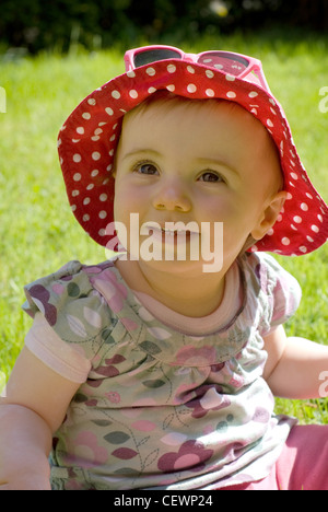 A female baby fair hair, wearing a floral t shirt, red polka dot summer hat and pink sunglasses on top, smiling, looking up Stock Photo