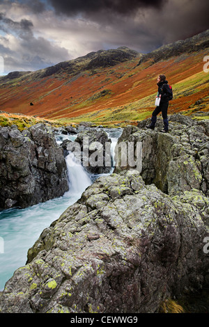 Young woman standing on boulders next to the River Esk. The River Esk begins in Great Moss below Scafell Pike in the English Lak Stock Photo