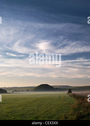 View across misty field to Silbury Hill, the tallest prehistoric human-made mound in Europe, beneath swirling clouds. Stock Photo