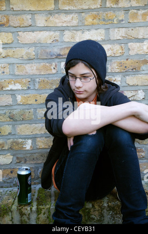 Teenage boy wearing black beanie orange t shirt and black hooded top and black jeans sitting on wall can of beer next to him Stock Photo