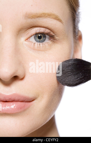 Female with fair hair off her face, wearing minimal make up, applying blusher to her cheek with a make up brush Stock Photo