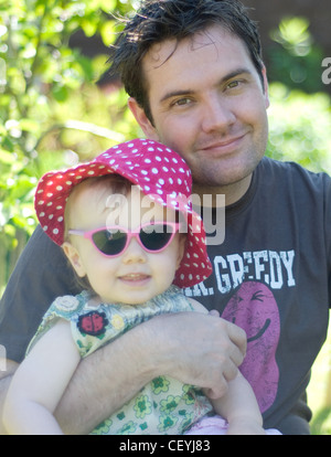 A female baby wearing a pink polka dot summer hat, green floral summer dress and pink sunglasses, sitting on lap of an adult Stock Photo