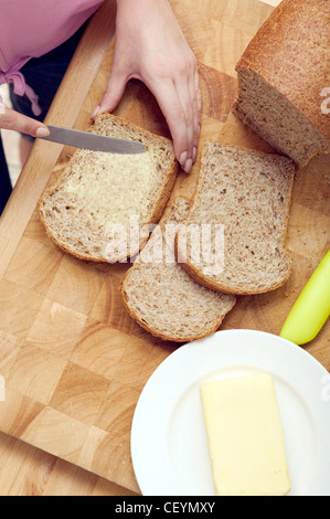 Female standing at kitchen work surface buttering slice of wholemeal bread on wood chopping board loaf of bread, carving knife Stock Photo