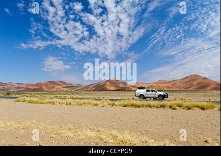 4x4 vehicle parked along the tarmac road in Sossusvlei with its red sand dunes,  Namib Naukluft Park. Namibia. Stock Photo