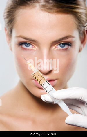 Female with blonde hair worn tied up wearing shimmery gold eye make up Botox syringe in front of her face Stock Photo